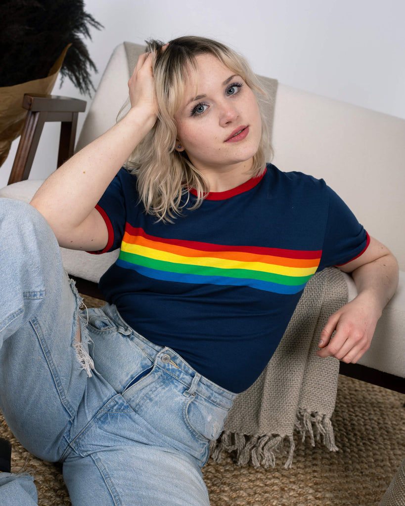 The model reclines casually in the Navy Ringer Retro Rainbow Striped T Shirt by Run & Fly, paired with light-wash ripped jeans. The bright rainbow stripe pops against the navy background, complemented by red ringer details. Surrounded by cosy home décor, this sustainable piece is perfect for retro enthusiasts.