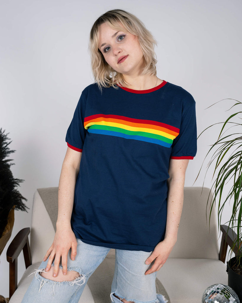 Front view of the Navy Ringer Retro Rainbow Striped T Shirt by Run & Fly, showcasing the vibrant rainbow stripe across the chest. Model wears it with light-wash distressed jeans, standing casually with a relaxed expression. The ethical, slow fashion tee combines bold retro vibes with sustainable style.