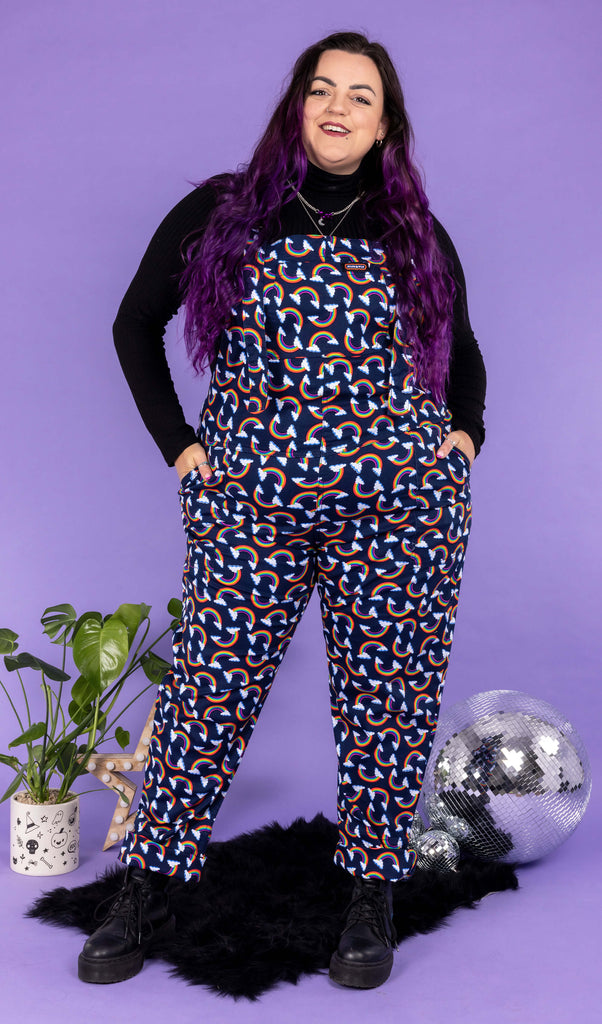 Luisa, a white femme model with purple hair, is wearing Over the Rainbow & Clouds Stretch Twill Dungarees with a long sleeve black turtle neck underneath and black boots. Luisa is stood in front of a lilac background on a black fluffy rug amongst disco balls, a star shaped light and a plant and is facing the camera smiling with her hands in the dungaree pockets.