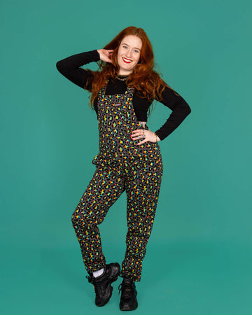 Isobella a femme model with long ginger hair is wearing Run & Fly Grey Rainbow Leopard Stretch Twill Dungarees paired with a cropped long sleeve black top and black trainers. The grey dungarees have an all over rainbow leopard print design. Isobella is smiling with one hand on her hip and other in her hair and one leg turned outwards in front of a green background.