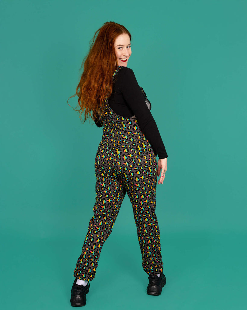 Isobella a femme model with long ginger hair is wearing Run & Fly Grey Rainbow Leopard Stretch Twill Dungarees paired with a cropped long sleeve black top and black trainers. The grey dungarees have an all over rainbow leopard print design. Isobella is stood with her back to the camera and is looking back over her shoulder and smiling at the camera in front of a green background. 