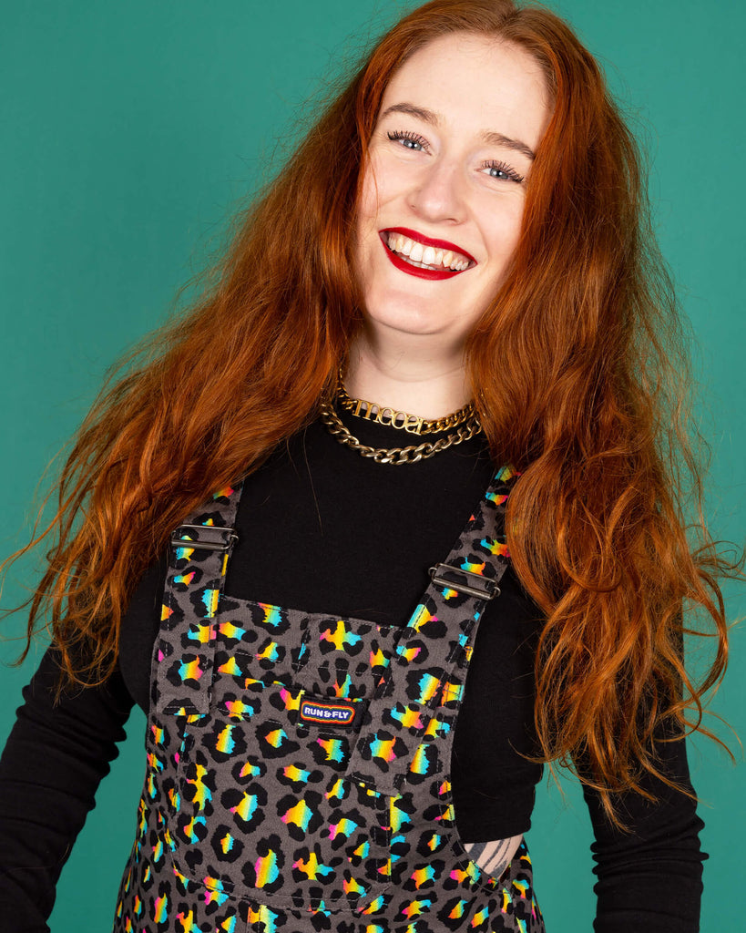 Isobella a femme model with long ginger hair is wearing Run & Fly Grey Rainbow Leopard Stretch Twill Dungarees paired with a cropped long sleeve black top. The grey dungarees have an all over rainbow leopard print design. Isobella is smiling at the camera with her hands in the pockets of the dungarees in front of a green background. 