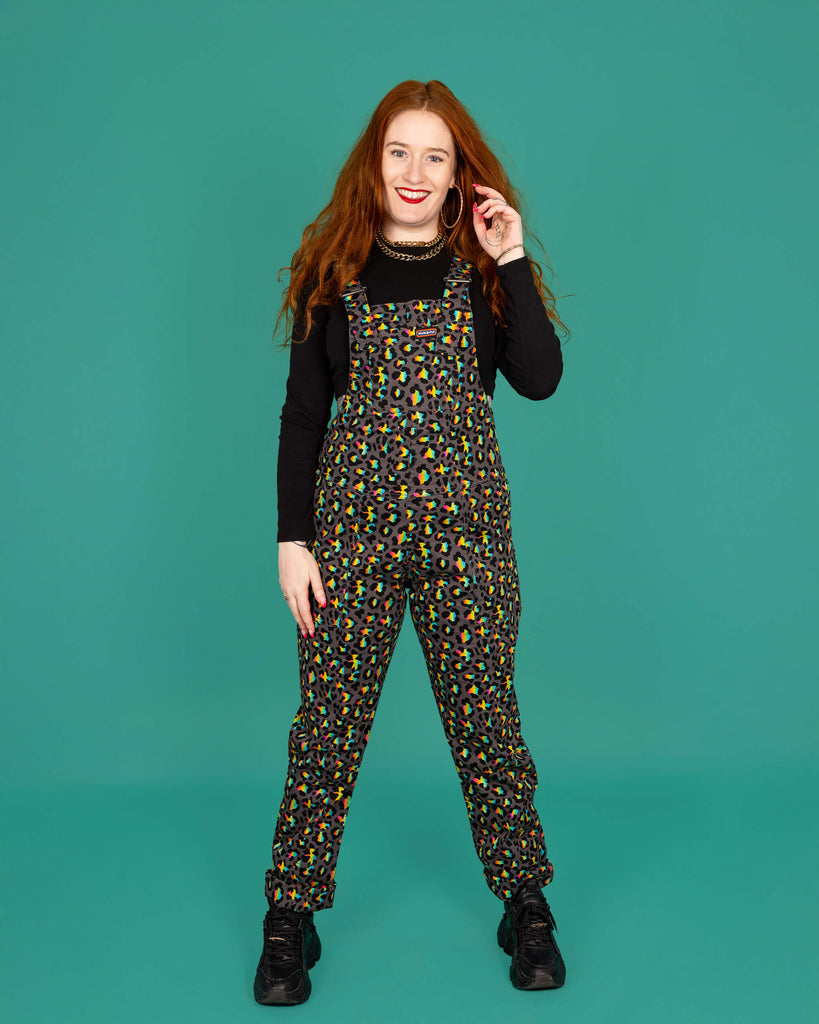 Isobella a femme model with long ginger hair is wearing Run & Fly Grey Rainbow Leopard Stretch Twill Dungarees paired with a cropped long sleeve black top and black trainers. The grey dungarees have an all over rainbow leopard print design. Isobella is stood facing the camera and smiling with one hand in her hair and other by her side in front of a green background. 