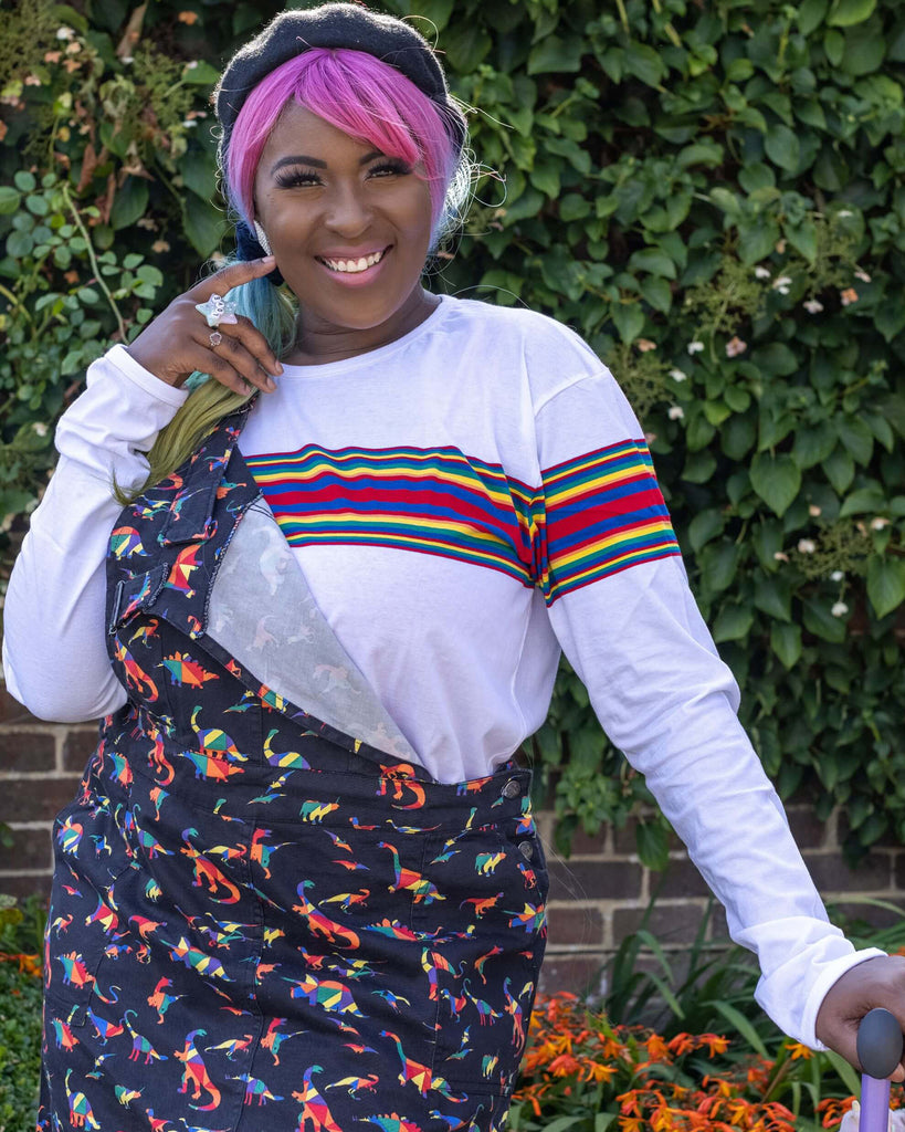 Ayesha smiling, holding a bright purple walking stick and wearing the Retro Rainbow Engineered Striped Long Sleeve T Shirt with a rainbow dino pinafore dress