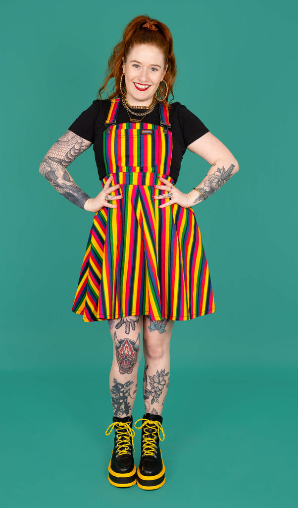 Isobela a model with long ginger hair and tattoos is wearing Rainbow Stripes Flared Pinafore Dress. The pinafore dress has vertical rainbow stripes all over. The pinafore has been paired with a short sleeve black top and boots. Isabella is stood facing the camera with her hands on her hips and smiling. The background of the photo is green.
