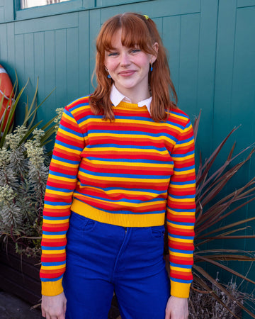 Tasha is wearing the Rainbow Primary Colours Striped Cropped Jumper with a white collared shirt underneath and blue jeans. She is stood outside in front of a blue wall with plants facing the camera and smiling with her arms down by her sides.