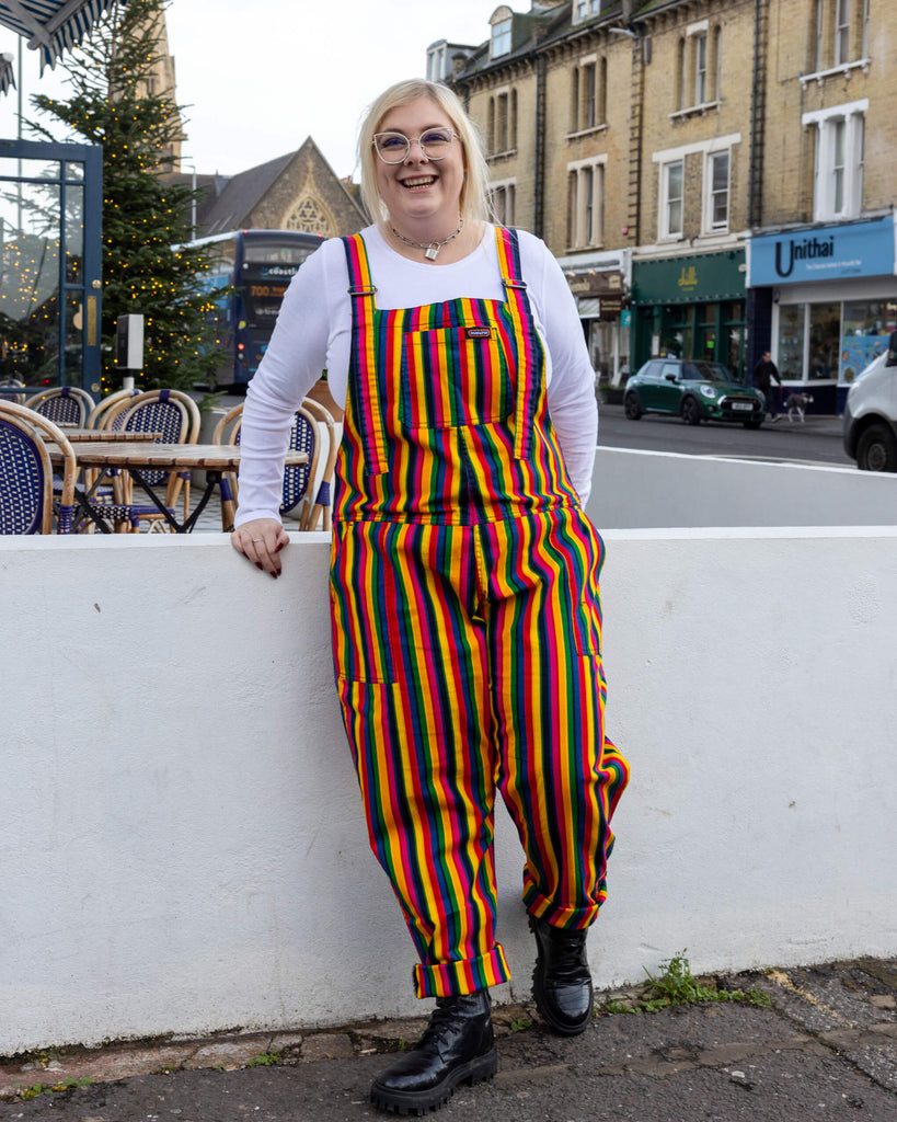 A smiling person wearing Run & Fly rainbow-striped dungarees, standing outdoors against a white wall. The dungarees feature vibrant vertical stripes in red, yellow, green, blue, and pink. They're paired with black combat boots and a white long-sleeved top. Run & Fly is a small ethical slow fashion brand specialising in bold, playful prints and eco-friendly fabrics.
