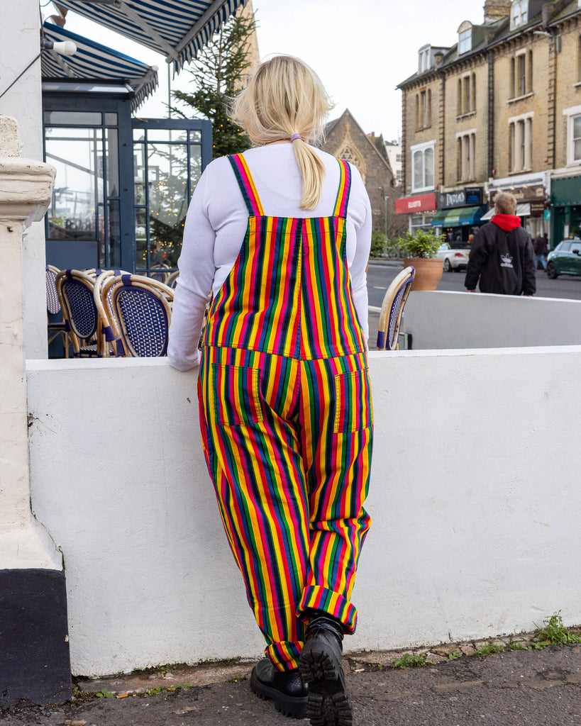 Back view of rainbow-striped dungarees from Run & Fly, featuring vibrant vertical stripes in red, yellow, green, blue, and pink. The person models the dungarees outdoors, leaning on a white wall. Paired with black boots and a white long-sleeved top. Run & Fly focuses on ethical slow fashion with unique, bold designs.