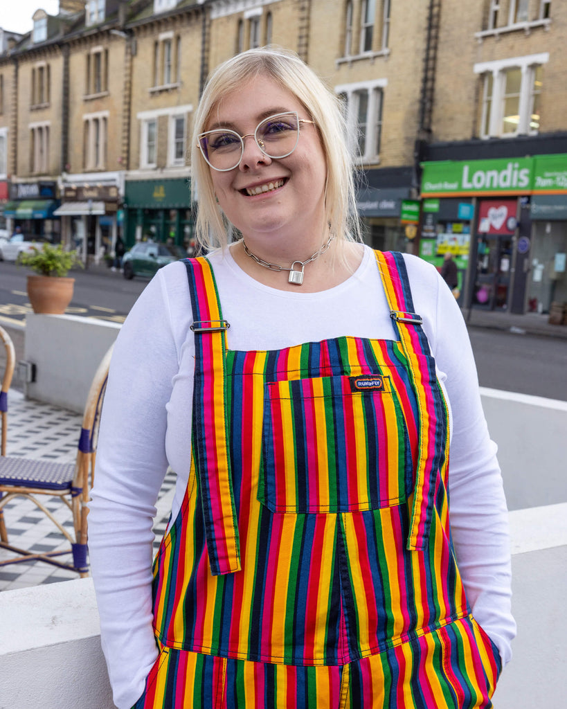 Close-up of a smiling person wearing Run & Fly rainbow-striped dungarees. The dungarees feature bold vertical stripes in bright, contrasting colours like red, yellow, green, blue, and pink. Styled with a white long-sleeved top and chunky silver necklace. Run & Fly creates fun, ethical, and sustainable clothing.