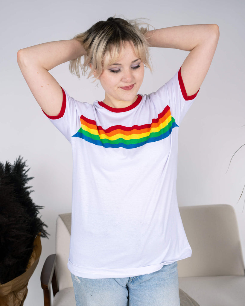 Model standing with arms playfully raised, showing off the Run & Fly White Ringer Retro Rainbow Striped T-Shirt. The vibrant rainbow stripe design pops against the white base, with red accents on the sleeves and neckline adding retro charm. Light-wash ripped jeans complete the cheerful, slow fashion look.