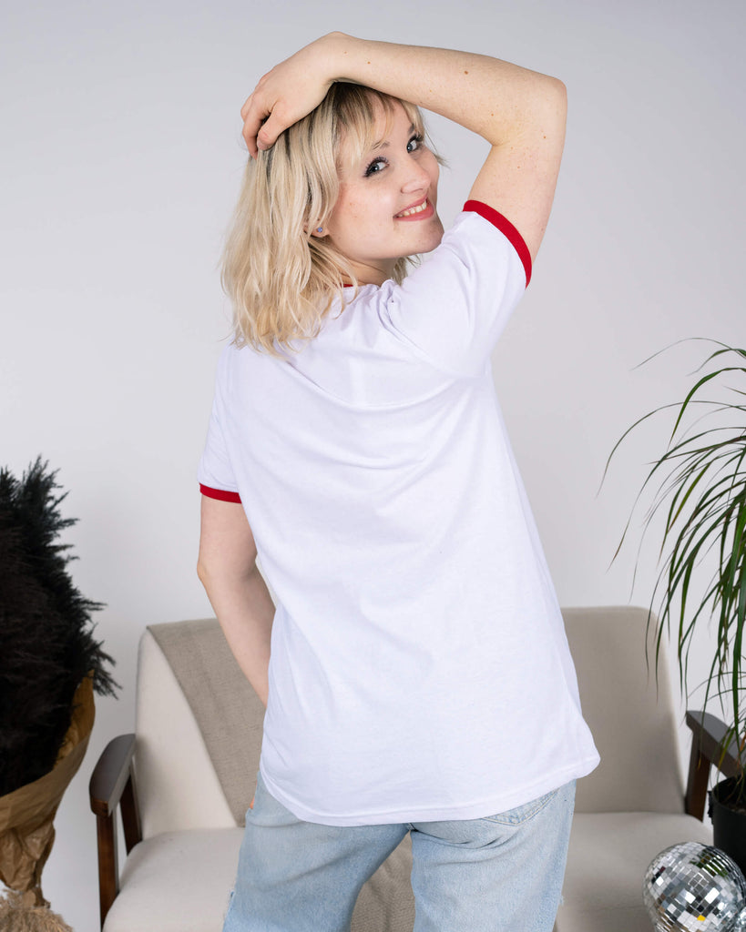 Model standing with their back turned, showcasing the plain white back of the Run & Fly White Ringer Retro Rainbow Striped T-Shirt. The red ringer detailing on the sleeves and neckline adds a retro flair. Styled with light-wash ripped jeans, the ethical, slow fashion tee is paired with a playful pose, perfect for everyday comfort and style.