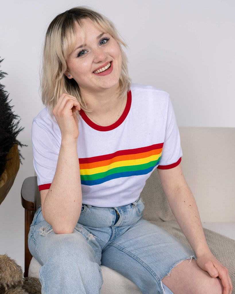 Model sitting on a beige chair, wearing the Run & Fly White Ringer Retro Rainbow Striped T-Shirt featuring bold rainbow stripes across the chest and red ringer detailing on the sleeves and neckline. Paired with light-wash ripped jeans, the look exudes cheerful retro vibes. The ethical slow fashion design is showcased with a friendly smile and relaxed pose, embodying fun and inclusivity.