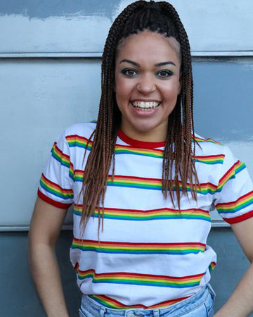 Model is smiling at the camera with hands in their light blue jean pockets, whilst wearing the Rainbow Block Striped T Shirt.