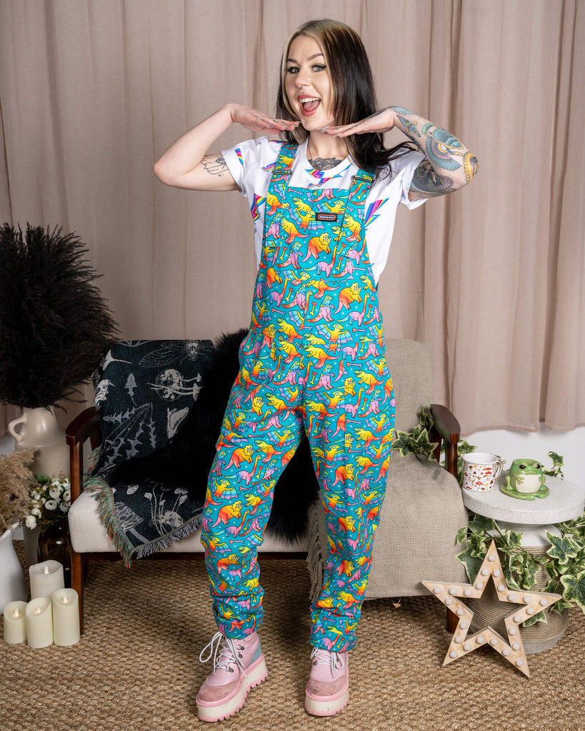 A tattooed model stands posing playfully in Run & Fly Rainbow Dinosaurs Stretch Twill Dungarees, smiling with hands under their chin. The dungarees have a fun rainbow dinosaur print by Waffle Mama, a relaxed fit, and five pockets. They’re styled with a white lightning bolt T-shirt and chunky pink platform boots. Ethically made with Oeko-Tex vegan dyes, perfect for dino lovers and quirky fashion fans.