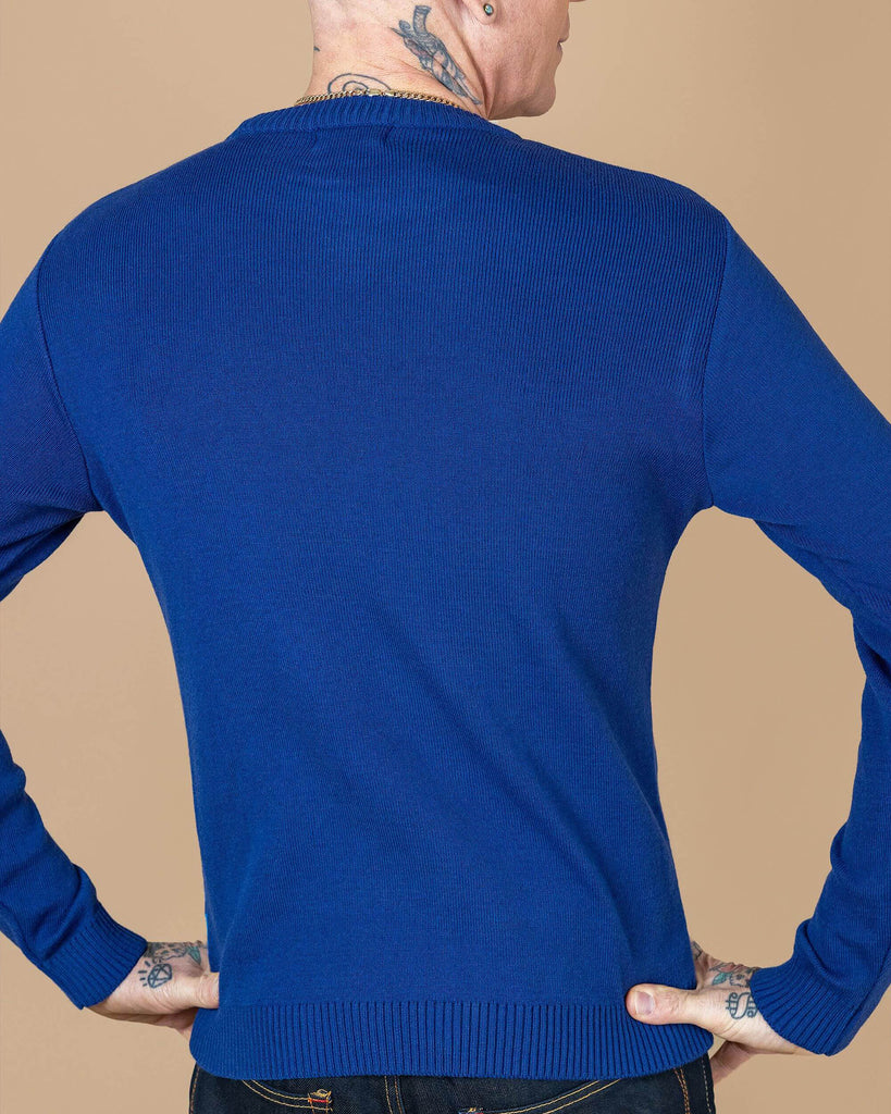 James is stood in front of a beige background wearing the Run and fly rainy days jumper with denim jeans. He is facing away from the camera with both hands resting on his hips and looking right. The jumper is a blue base with a red, orange, white and blue stripe section, a stripe of rain clouds, a stripe of red umbrellas and a stripe of light blue welly boots. The back and sleeves are a plain blue. The photo is cropped from the hips up.