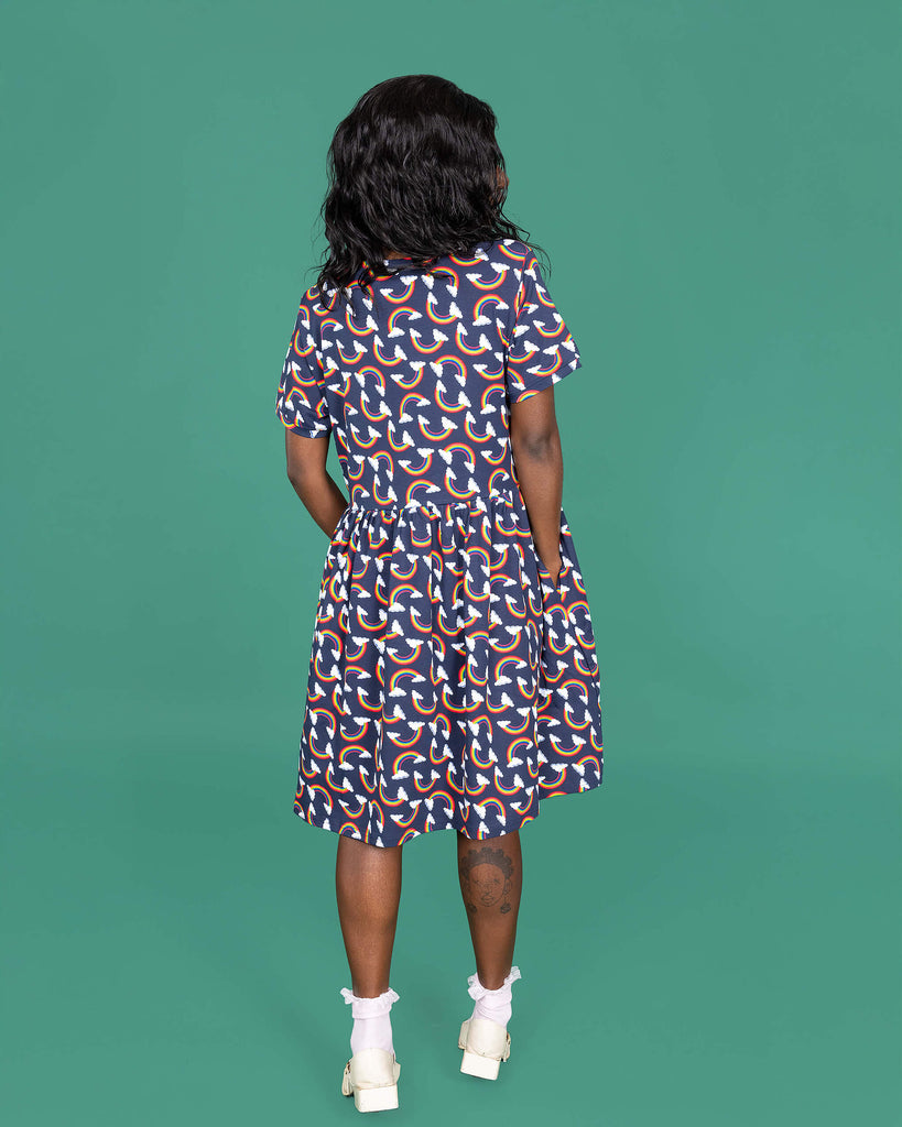 The Over the Rainbow & Clouds Oversized Smock Dress worn by a femme alternative model with black hair with white frilly socks and white dolly shoes on a teal studio background. She is facing away from camera looking to the right with both hands resting in the dress side pockets. The navy blue base dress has repeating rainbow arches with white clouds.