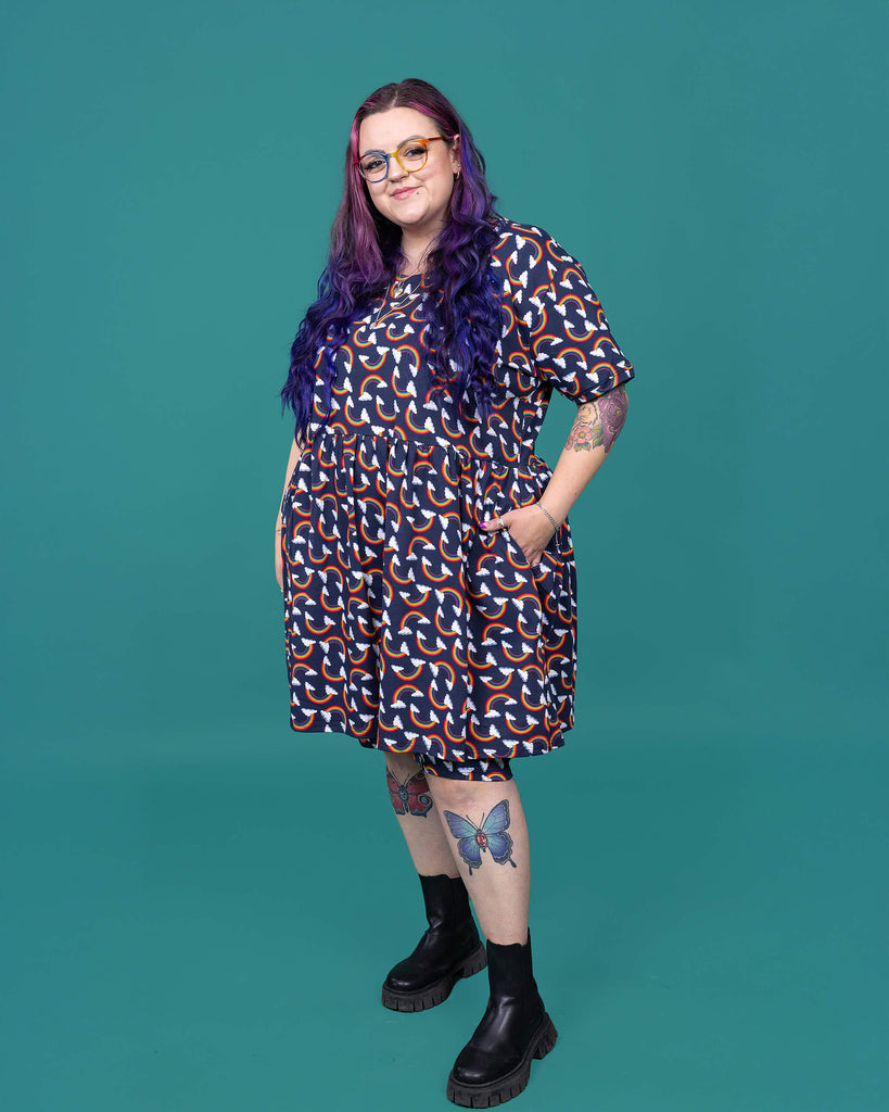 The Over the Rainbow & Clouds Oversized Smock Dress worn by a femme alternative tattooed model with rainbow glasses, long pink to purple hair, the matching cycle shorts and black boots on a teal studio background. She is facing forward angled left smiling with both hands resting in the dress pockets. The navy blue base dress has repeating rainbow arches with white clouds.