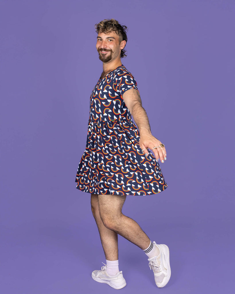 The Over the Rainbow & Clouds Oversized Smock Dress worn by a masc non binary person with a short curly mullet, matching cycle shorts and white trainers on a purple studio background. They are facing left laughing whilst swishing the dress and twirling. The navy blue base dress has repeating rainbow arches with white clouds.
