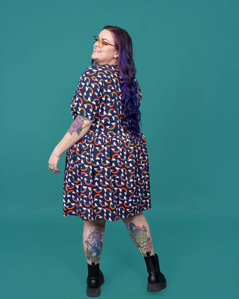 The Over the Rainbow & Clouds Oversized Smock Dress worn by a femme alternative tattooed model with rainbow glasses, long pink to purple hair, the matching cycle shorts and black boots on a teal studio background. She is facing away from camera laughing looking over her left shoulder with her arms out to the side. The navy blue base dress has repeating rainbow arches with white clouds.