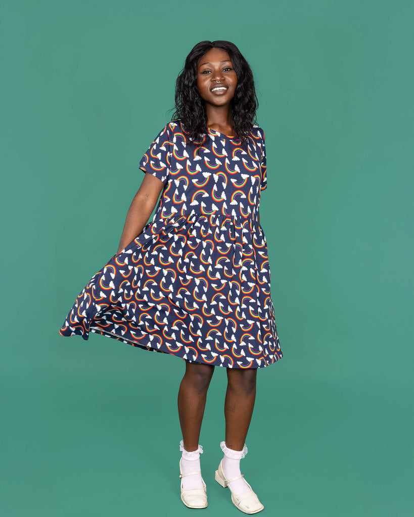 The Over the Rainbow & Clouds Oversized Smock Dress worn by a femme alternative model with black hair with white frilly socks and white dolly shoes on a teal studio background. She is facing forward swishing the dress with both hands in the pockets and smiling to camera. The navy blue base dress has repeating rainbow arches with white clouds.