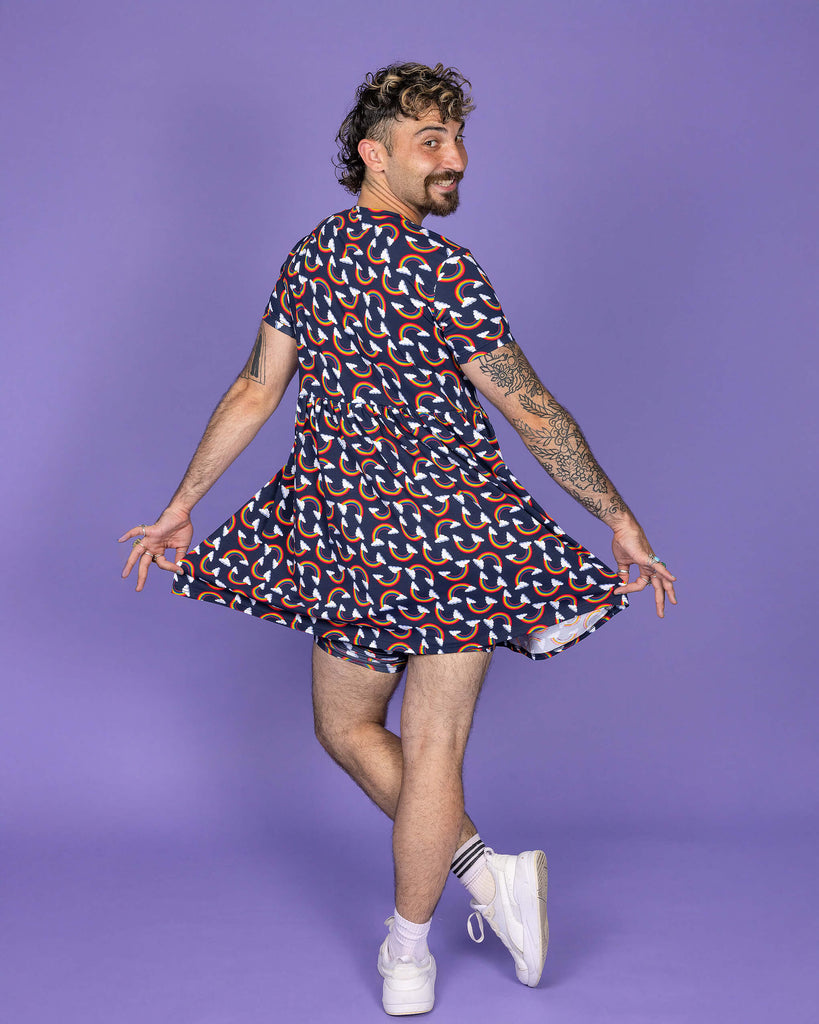 The Over the Rainbow & Clouds Oversized Smock Dress worn by a masc non binary person with a short curly mullet, matching cycle shorts and white trainers on a purple studio background. They are facing away holding out the bottom of the dress and crossing one leg over the other smiling looking over their right shoulder to camera. The navy blue base dress has repeating rainbow arches with white clouds.