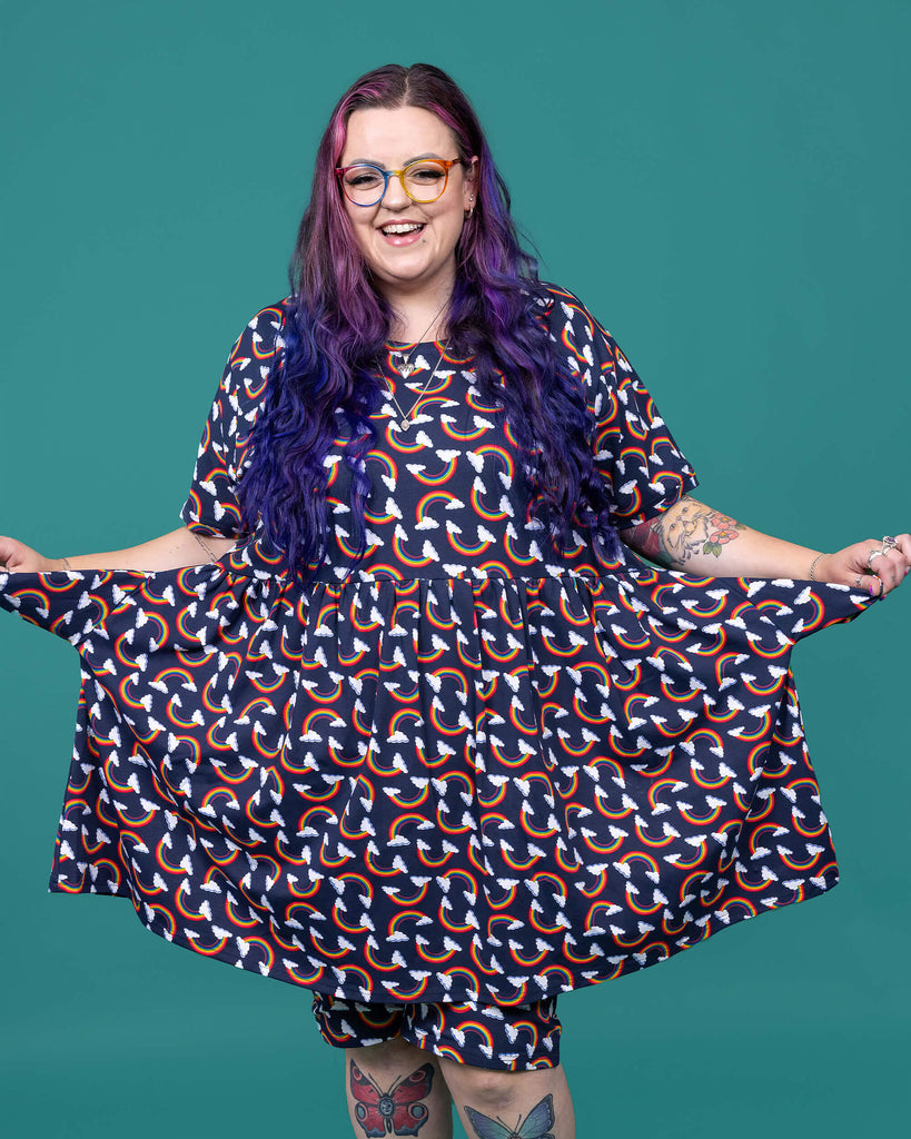 The Over the Rainbow & Clouds Oversized Smock Dress worn by a femme alternative tattooed model with rainbow glasses, long pink to purple hair, the matching cycle shorts and black boots on a teal studio background. She is facing forward laughing holding out the smock dress pockets to the side.The navy blue base dress has repeating rainbow arches with white clouds. The navy blue base dress has repeating rainbow arches with white clouds.