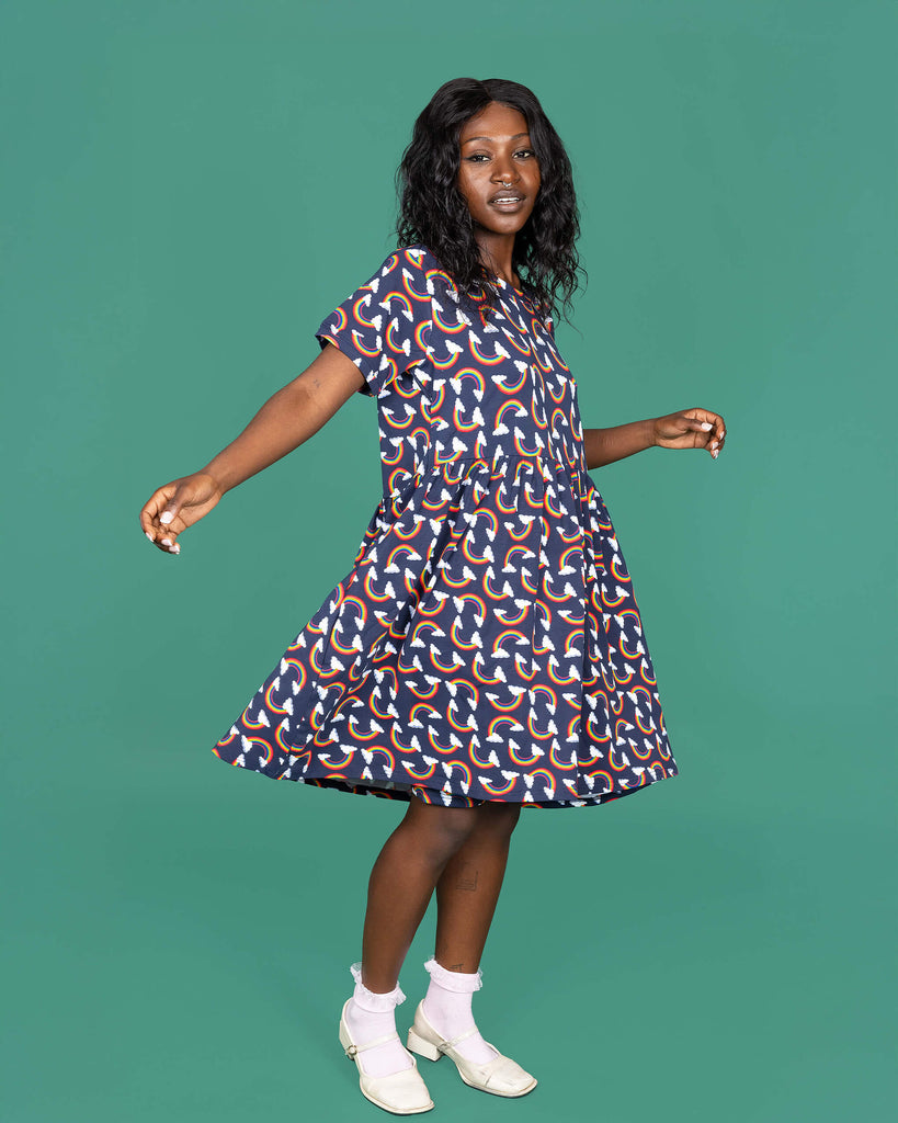 The Over the Rainbow & Clouds Oversized Smock Dress worn by a femme alternative model with black hair with white frilly socks and white dolly shoes on a teal studio background. She is facing forward swishing the dress out with both hands out to the side whilst smising to camera. The navy blue base dress has repeating rainbow arches with white clouds.