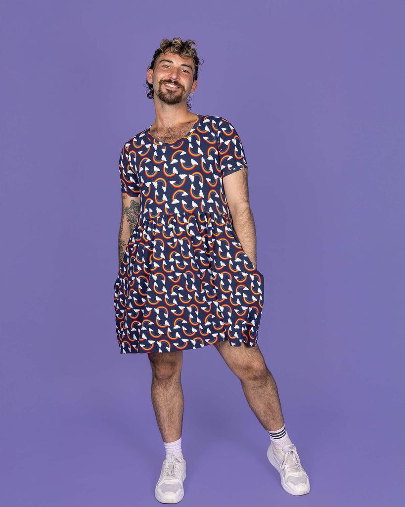 The Over the Rainbow & Clouds Oversized Smock Dress worn by a masc non binary person with a short curly mullet, matching cycle shorts and white trainers on a purple studio background. They are facing forward laughing leaning more on their left hip with both hands resting in the dress pockets. The navy blue base dress has repeating rainbow arches with white clouds.