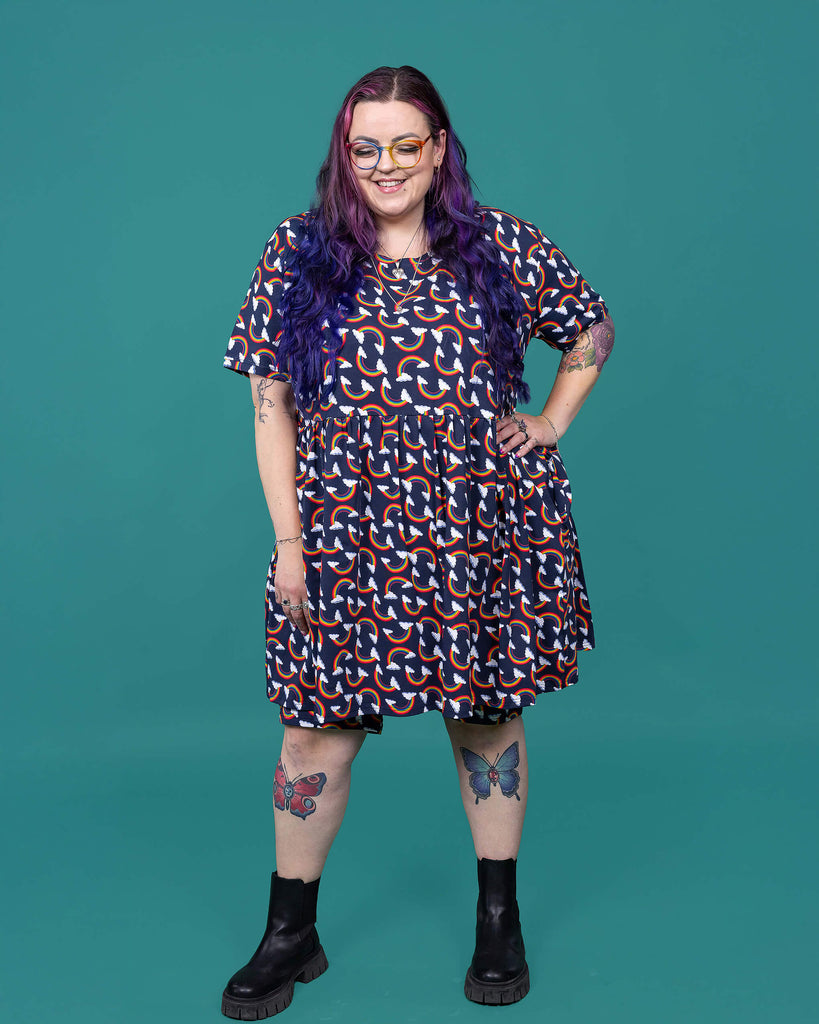 The Over the Rainbow & Clouds Oversized Smock Dress worn by a femme alternative tattooed model with rainbow glasses, long pink to purple hair, the matching cycle shorts and black boots on a teal studio background. She is facing forward laughing with her eyes closed and her hand on her hip. The navy blue base dress has repeating rainbow arches with white clouds.
