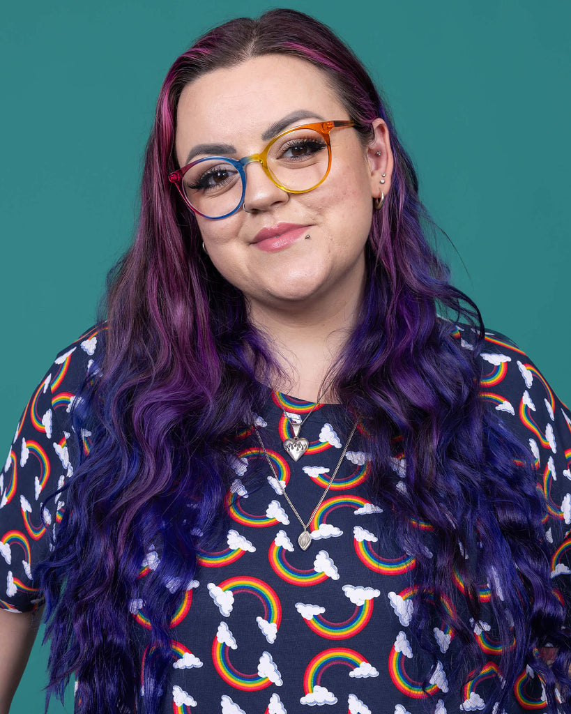 Close up of the Over the Rainbow & Clouds Oversized Smock Dress worn by a femme alternative tattooed model with rainbow glasses and long pink to purple hair on a teal studio background. She is facing forward smiling holding out the skirt of the dress. The navy blue base dress has repeating rainbow arches with white clouds.