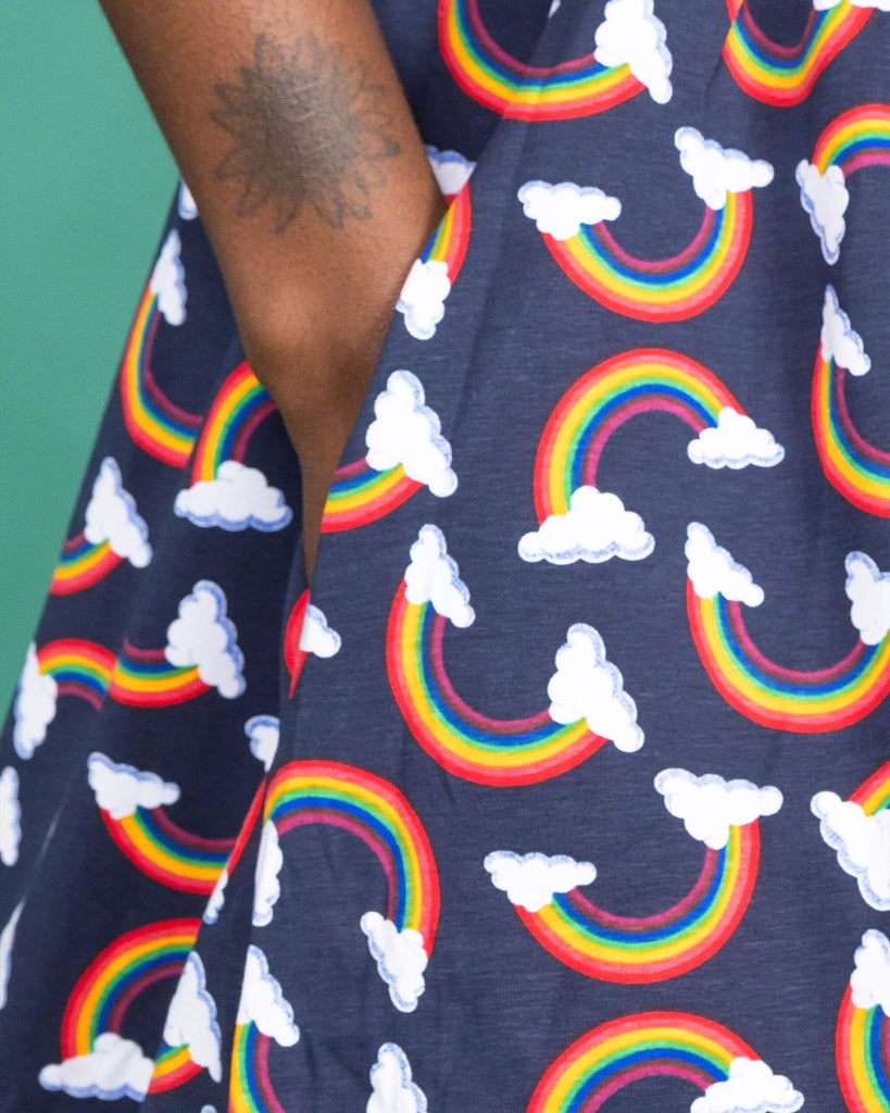 Close up of the Over the Rainbow & Clouds Oversized Smock Dress worn by a femme alternative model on a teal studio background. She is facing right with her hand in the side pocket. The navy blue base dress has repeating rainbow arches with white clouds.