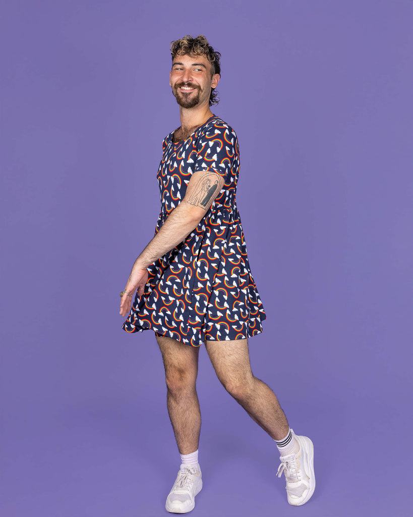 The Over the Rainbow & Clouds Oversized Smock Dress worn by a masc non binary person with a short curly mullet, matching cycle shorts and white trainers on a purple studio background. They are facing forward laughing whilst swishing the dress and taking a step. The navy blue base dress has repeating rainbow arches with white clouds.