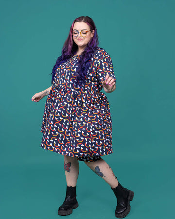 The Over the Rainbow & Clouds Oversized Smock Dress worn by a femme alternative tattooed model with rainbow glasses, long pink to purple hair, the matching cycle shorts and black boots on a teal studio background. She is facing forward taking a step to the left laughing with her eyes closed and swishing the dress. The navy blue base dress has repeating rainbow arches with white clouds.