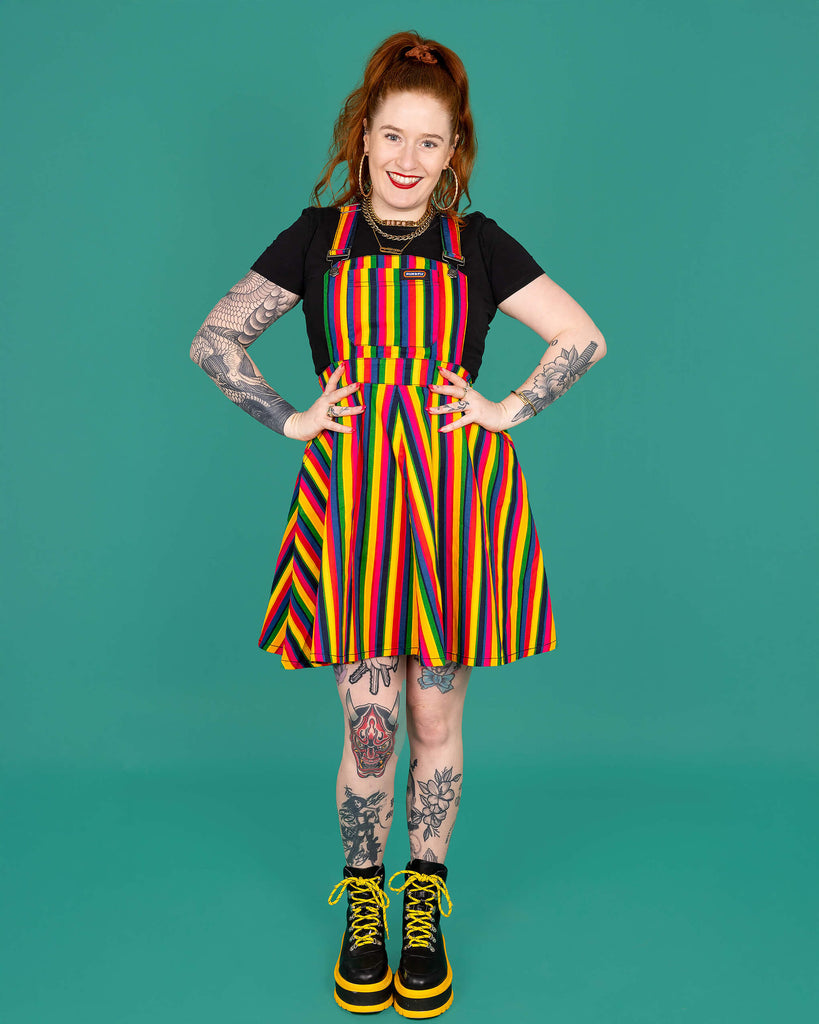 Isobela a model with long ginger hair and tattoos is wearing Rainbow Stripes Flared Pinafore Dress. The pinafore dress has vertical rainbow stripes all over. The pinafore has been paired with a short sleeve black top and boots. Isabella is stood facing the camera with her hands on her hips and smiling. The background of the photo is green.