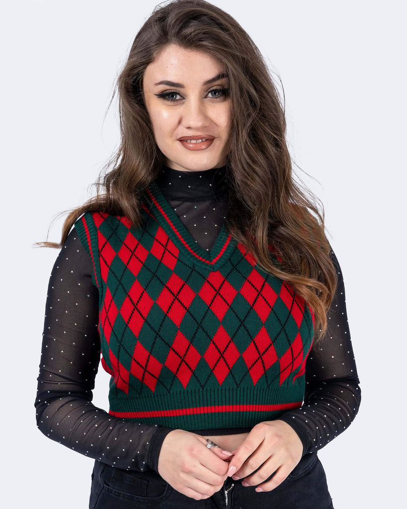 Charlotte is wearing Red and Green Argyle Knitted Tank Top with a long sleeve black mesh top underneath with white polka dots and black trousers. Charlotte is stood in front of a white background facing the camera smiling with her hands together. The photo is cropped from the thighs down. 