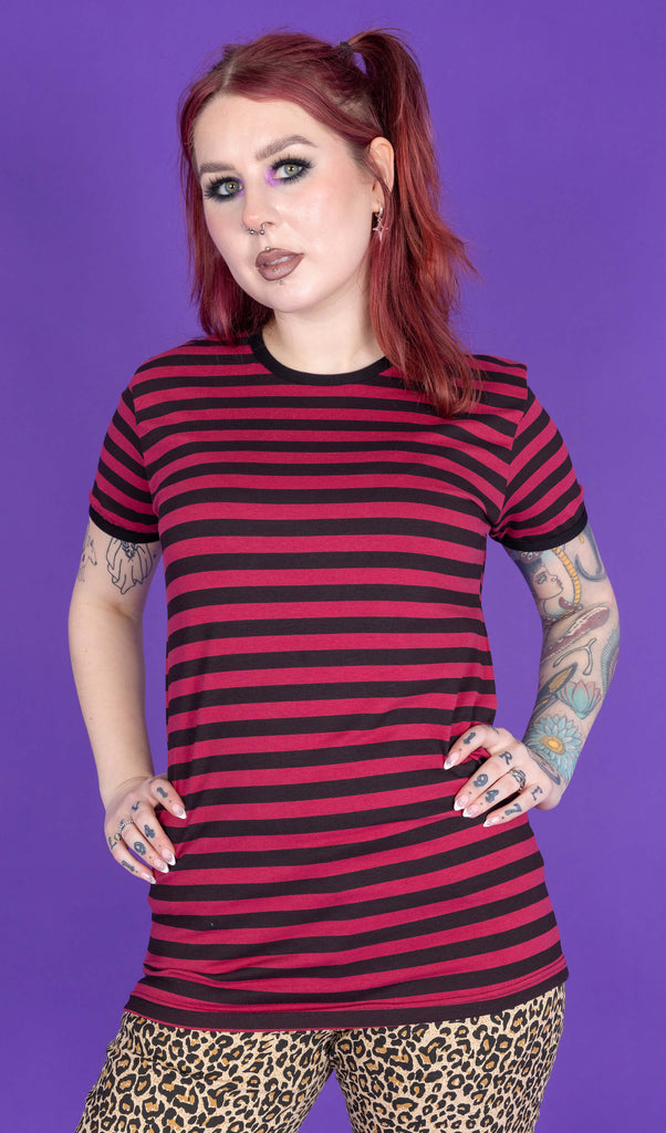 tattooed model with red hair wearing a black and red striped short sleeve top, paired with leopard print jeans. The model is posing with her hands on her hips in front of a purple background.