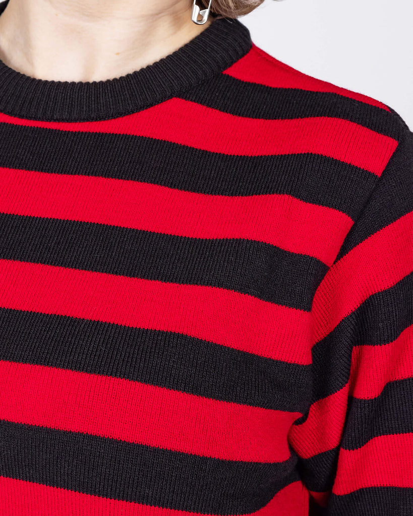 Close up of the Red and Black Striped Jumper worn by a femme model with short blonde hair with black trousers. She is facing forward to highlight the black neck and sleeve trim with alternating black and red stripes all over.