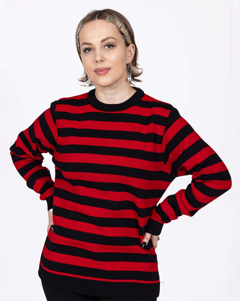 The Red and Black Striped Jumper worn by a femme model with short blonde hair with black trousers. She is facing forward smiling with her hands on her hips. The jumper has a black neck and sleeve trim with alternating black and red stripes all over.