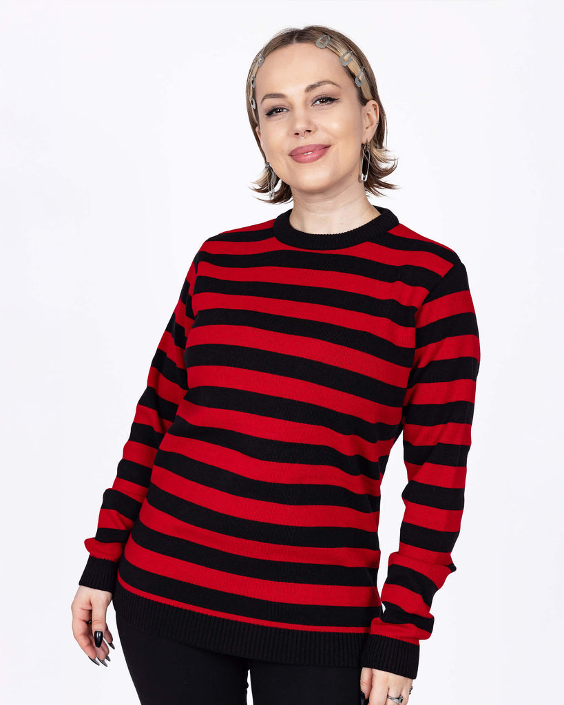 The Red and Black Striped Jumper worn by a femme model with short blonde hair with black trousers. She is facing forward smiling with her hands resting by her side. The jumper has a black neck and sleeve trim with alternating black and red stripes all over.