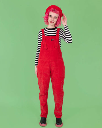 A pink haired femme model wearing the run and fly red stretch corduroy dungarees with a red beret, black boots and a black and white stripe long sleeve tshirt. They are facing forward to highlight the retro roller straps and front bib pocket with a rainbow run & fly label. They are smiling to camera with one hand resting by their side whilst the other rests on the beret.