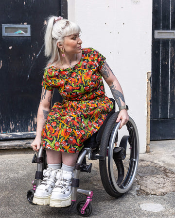 Model posing in a wheelchair looking off to one side outdoors on a sunny day wearing Run & Fly Red Panda belted tea dress with pockets. The print is filled with adorable red pandas surrounded by green leaves and orange branches on a black background. 
