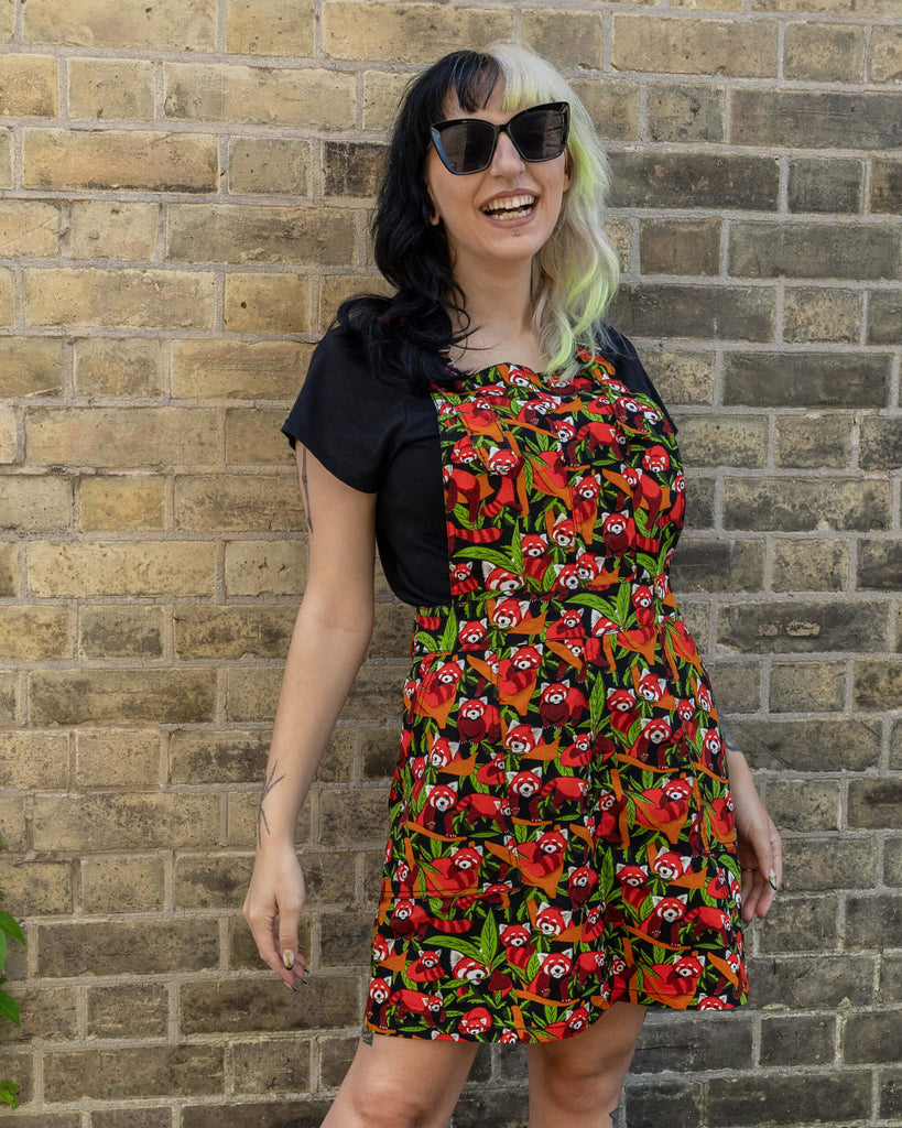A full view of a woman with black and blonde hair wearing a Run & Fly Red Panda pinafore. The pinafore features a lively red panda and green foliage pattern on a black background. She is posing playfully with a smile, her hands on her hips.