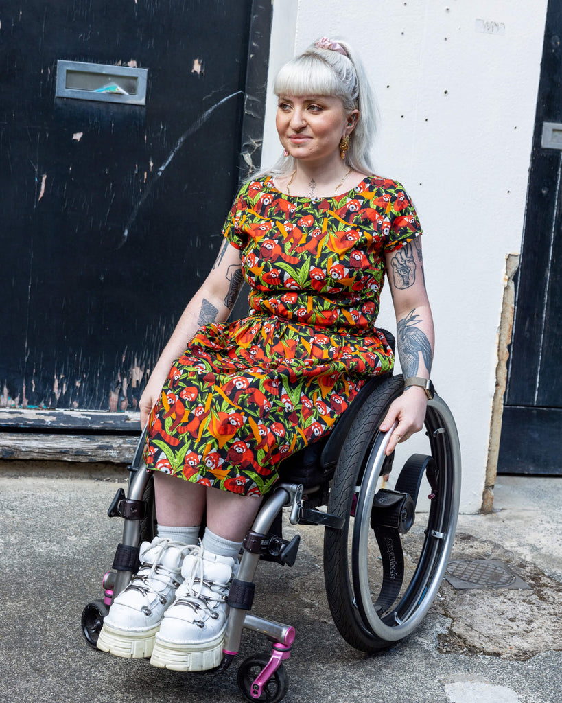 Female model posing in a wheelchair smiling at the camera, wearing Run & Fly Red Panda belted tea dress with pockets. The print has illustrations of red pandas among green leaves and orange foliage on a dark backdrop