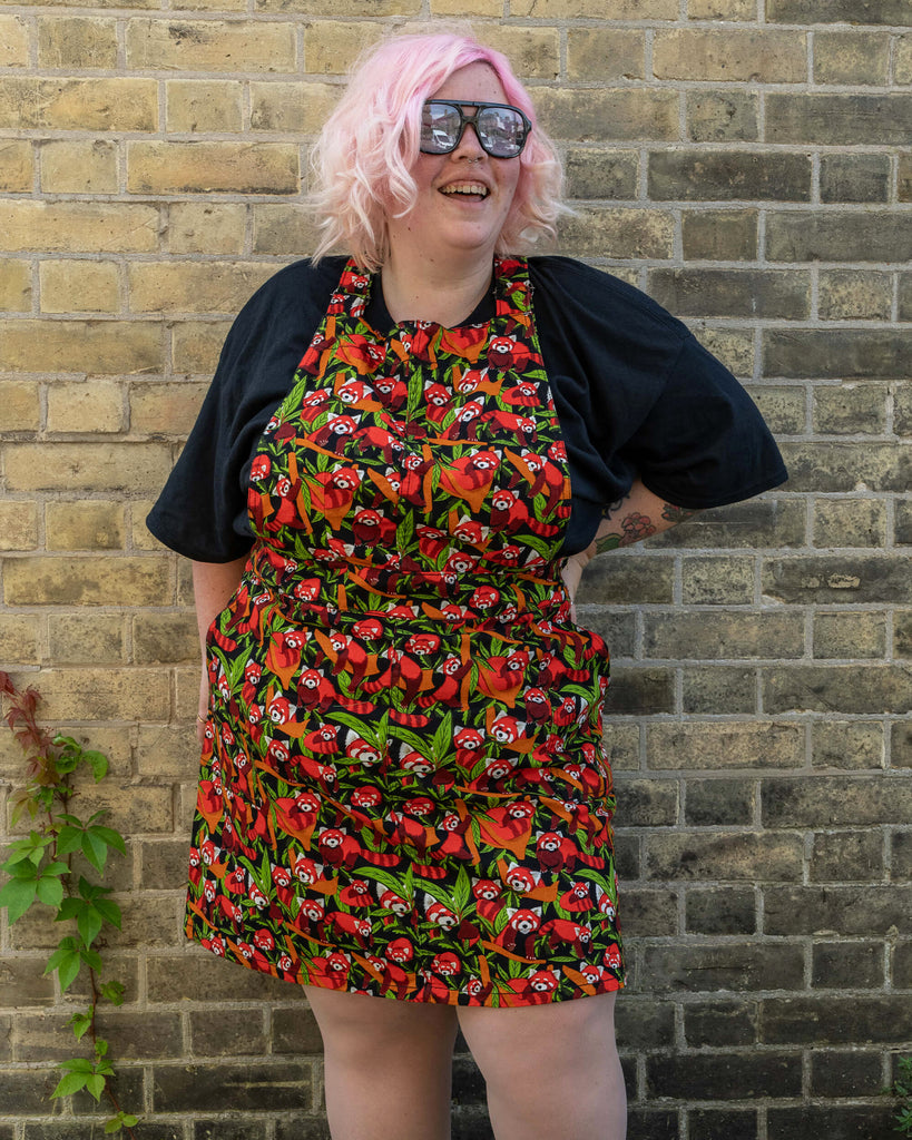 A model with pink hair and glasses is modelling a Run & Fly Red Panda pinafore. The pinafore displays a colourful pattern of red pandas and green leaves on a black background. They are standing in front of a brick wall, smiling, and has their hands in the pockets of the pinafore.