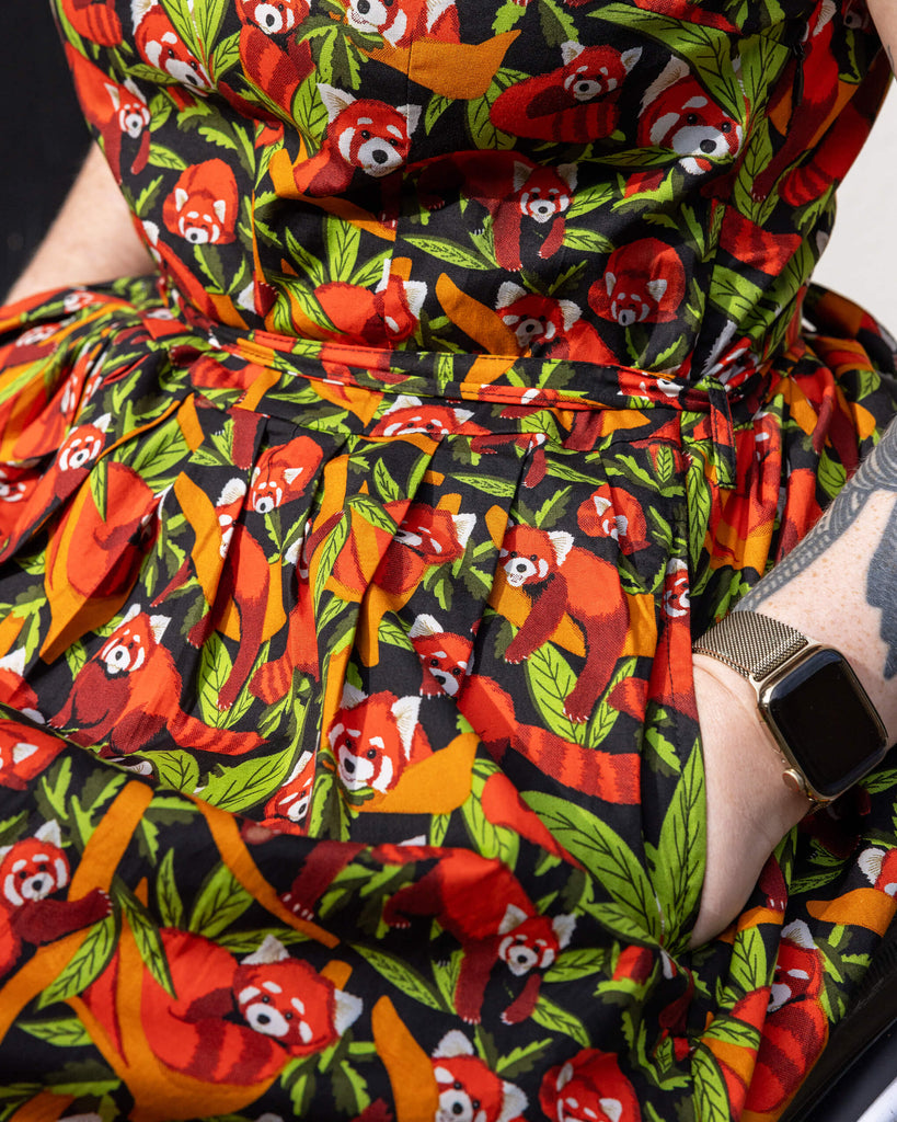 Close-up of Run & Fly Red Panda belted tea dress with pockets worn by a model in a wheelchair The dress has an all over print of red pandas surrounded by foliage, creating a striking contrast on a dark background. The model's hands are resting in the pockets of the dress.