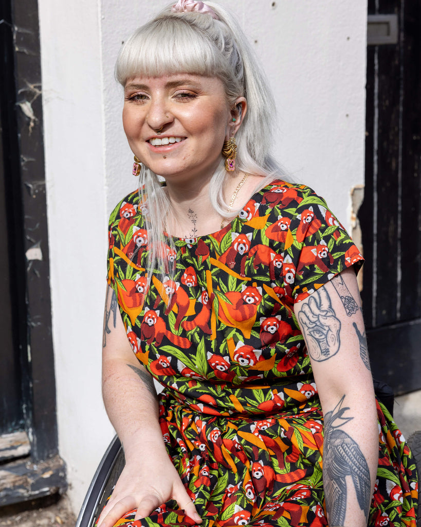 Model posing in a wheelchair smiling in the sun, wearing Run & Fly Red Panda belted tea dress with pockets. The print is filled with charming red pandas among green and orange foliage, set against a deep black background with a matching fabric waist belt. 