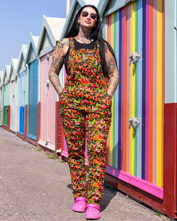 The Red Pandas Stretch Twill Dungarees worn by a femme tattooed model with long black hair, a black tank top, sunglasses and pink croc shoes. She is stood outside in the sunshine posing in front of colourful beach huts along the seafront. She is facing forward smiling with her hand on her hip and the other hand resting up on the beach hut. The black base print features various illustrated red pandas sleeping, eating and curled up on bamboo.