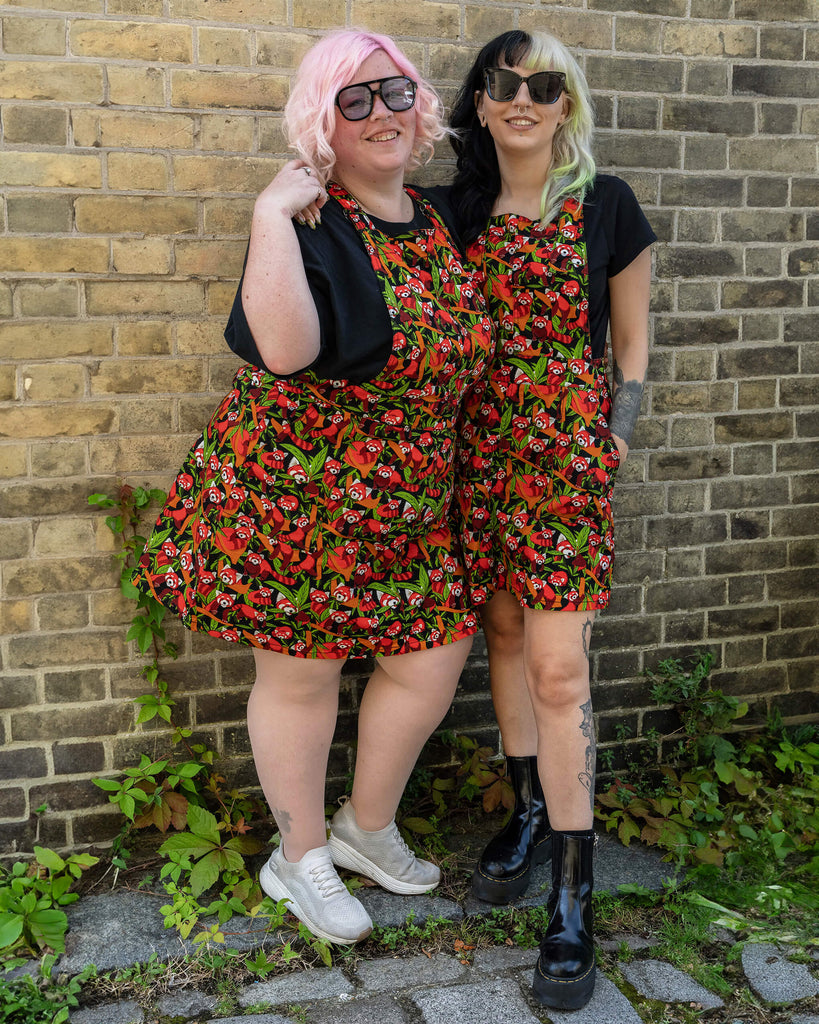 Two models are standing together, both wearing Run & Fly Red Panda pinafores. The pinafores feature a vibrant red panda and foliage pattern on a black background. One model has pink hair and is wearing glasses, while the other has black and blonde hair. Both are wearing black tops underneath their pinafores.