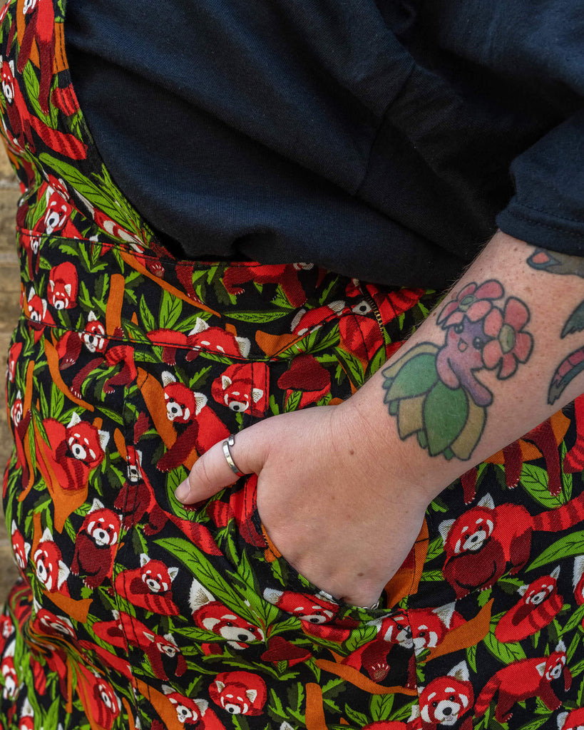 A close-up of the pockets on the Run & Fly Red Panda pinafore. The fabric features a colourful red panda and foliage design on a black background. The model’s hand is partially in the pocket, showcasing the detailed print and practicality of the design.