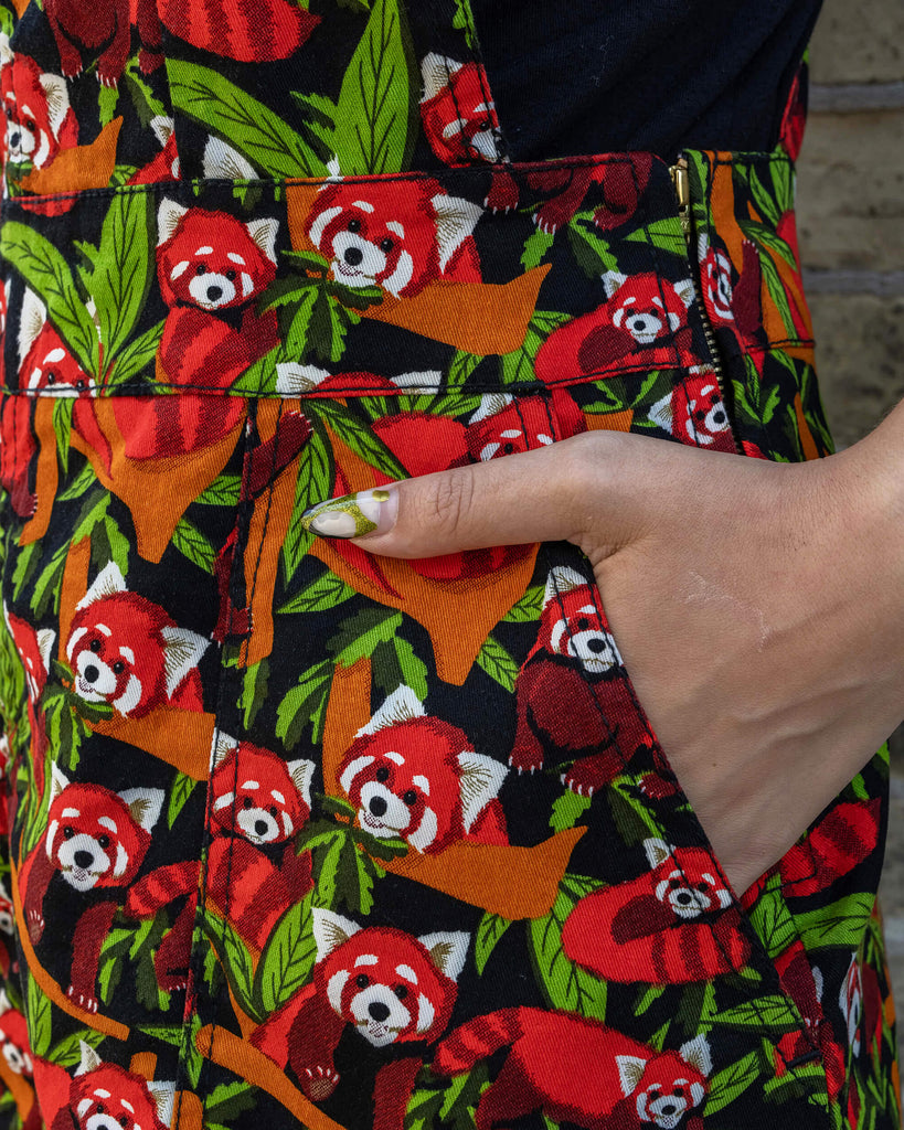 A close-up of the pockets on the Run & Fly Red Panda pinafore. The fabric features a colourful red panda and foliage design on a black background. The model’s hand is partially in the pocket, showcasing the detailed print and practicality of the design.